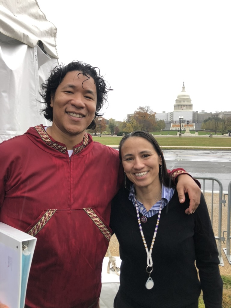 A person in a red jacket with their arm around a person in a black sweater. Both are standing outside with the US Capitol in the background.