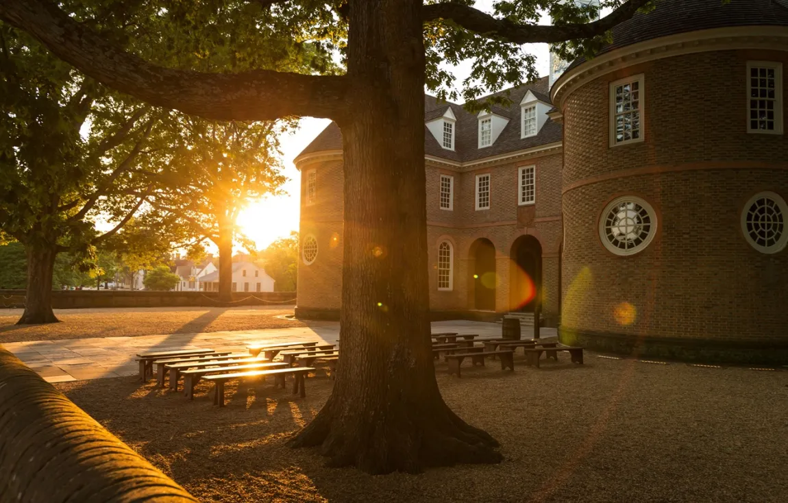 Sun setting behind trees and brick building, with benches and tree in foreground in Colonial Williamsburg